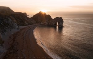 Durdle Door as the sun rises, and a lone admirer stands as a tiny figure beneath the huge rock